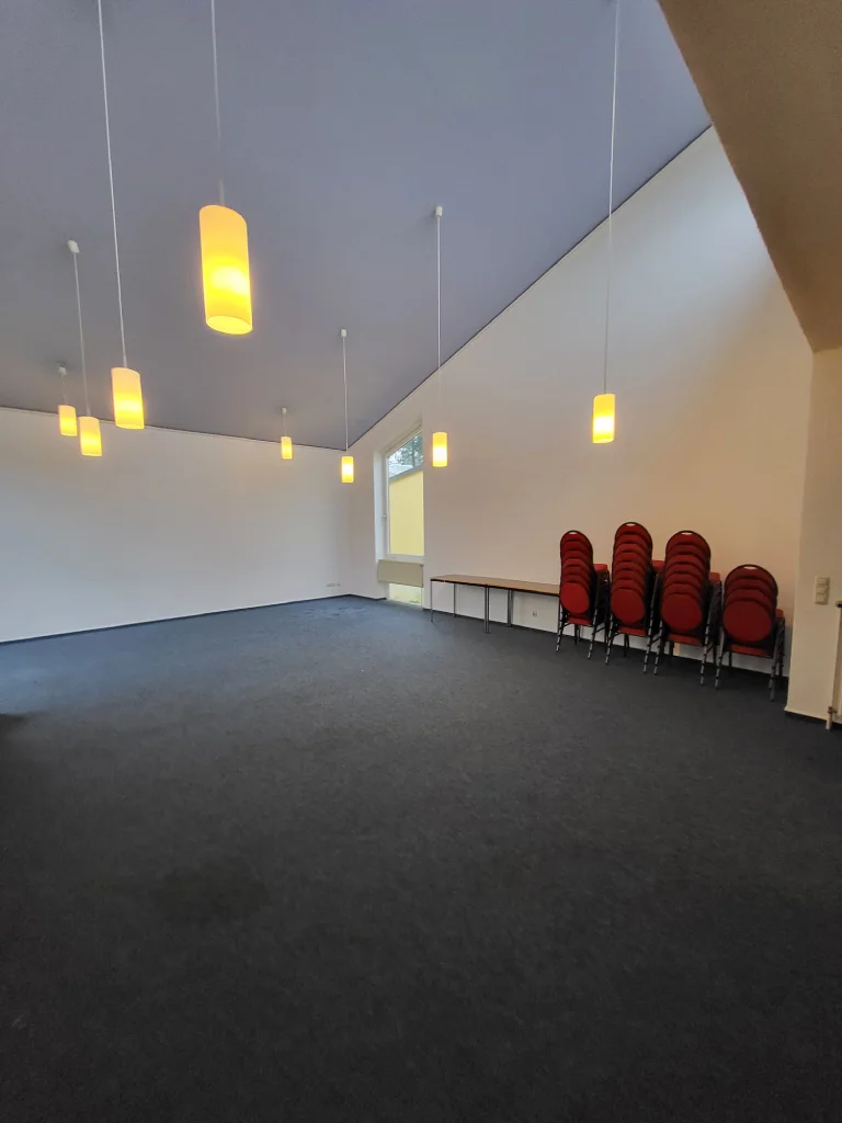 Heller Raum mit Deckenlampen und gestapelten Stühlen an der Wand – Evangelische Gesellschaft für Deutschland KdöR